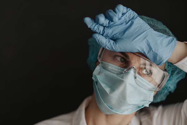 image of an essential worker in protective mask and gloves resting her hand against her forehead.